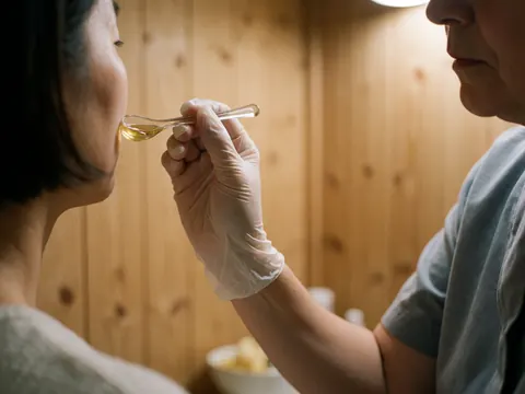 A professional aesthetician uses a spoon to apply honey treatment to a client's face in a salon with wooden walls and warm lighting.