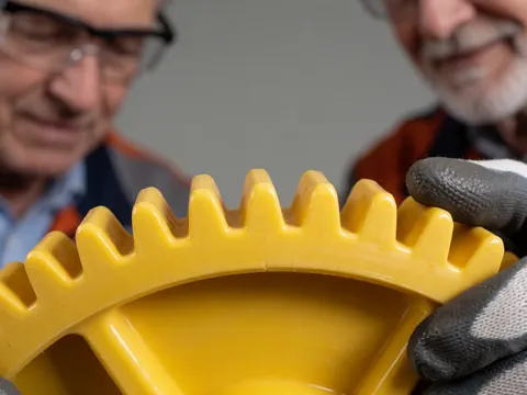Close-up of a gloved hand holding a large yellow gear in a factory setting with blurred workers in safety glasses in the background.