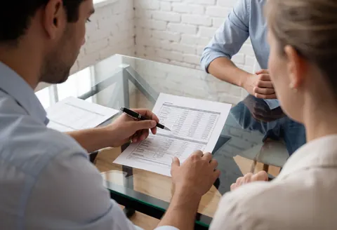 Two professionals review financial documents on a glass table in a modern office with a white brick wall.