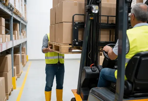 Worker using a forklift to load a pallet in a warehouse with high shelves and safety vests visible.