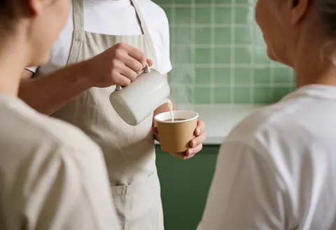 A barista pours steaming milk into a glass of hot cocoa in a kitchen with green tiles while two customers stand nearby.