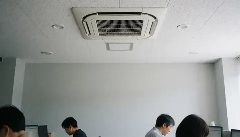 Modern office ceiling with cassette air conditioner, people working at computers, gray walls, natural light.