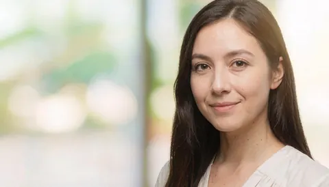 A smiling woman wearing a white shirt with a softly blurred background featuring gentle lights, suitable for corporate headshots.