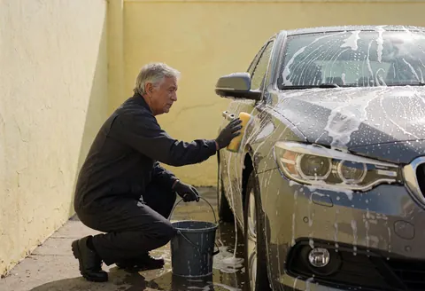 A man washes a car using a sponge and bucket in a paved area with a yellow wall, demonstrating an active car wash service.