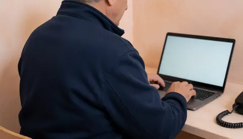 Adult man in dark jacket working on a laptop at a light wood desk in a modern office with terracotta walls.