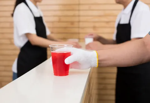 A hand wearing a white glove places a plastic cup with a red beverage on a fast food counter with light wood walls.