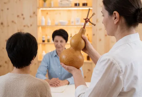 A practitioner holds a gourd with sticks while attending to a seated client in a room with wooden shelves and walls.