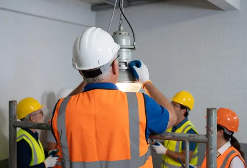 An electrician adjusts an industrial light fixture while standing on metal scaffolding inside a warehouse.