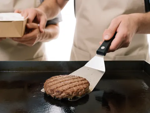 A cook in an apron uses a spatula to serve a hamburger from a steel griddle with visible steam in a fast food restaurant.