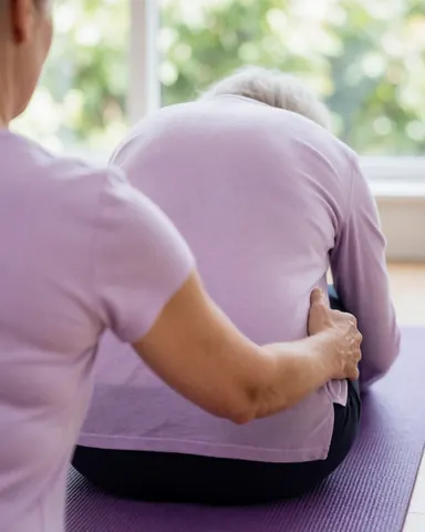 A yoga instructor assists an older woman in a forward fold posture on a purple mat near a window in a studio.