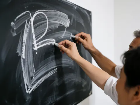 Close-up of an adult hand writing with white chalk on a blackboard, showing detailed handwriting for online course instruction.
