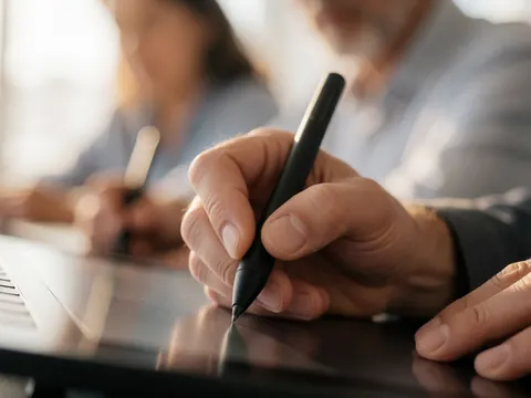 Close-up of an adult hand using a stylus to draw on a digital tablet with a blurred background of people working on graphic design.