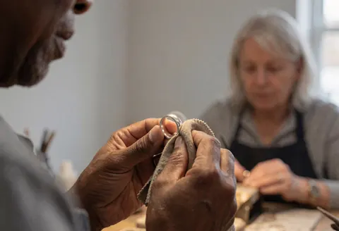 A close-up view of a hand using a soft cloth to polish a silver ring in a workshop setting with natural light and gray walls.