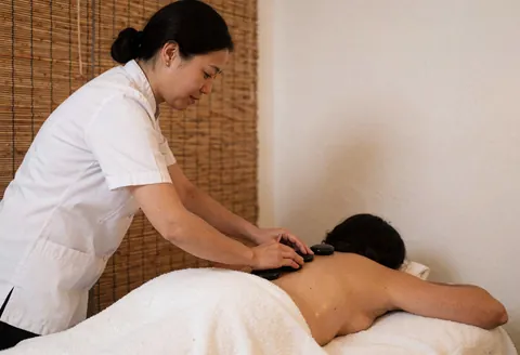 Therapist in white uniform applying hot stones to a client lying on a white towel in a spa with bamboo blinds