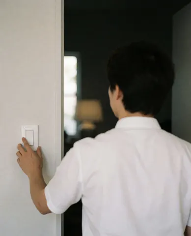 A professional in a white shirt adjusts a light switch on a clean wall with dim lighting in the background.