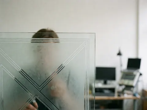Close-up of geometric patterns etched into glass, a hand holding an engraving tool, blurred workshop background with white walls