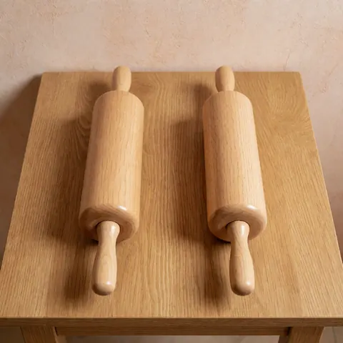 Two polished wooden rolling pins resting on a light wood table with a terracotta plaster wall, ideal for bakery and pastry product photography