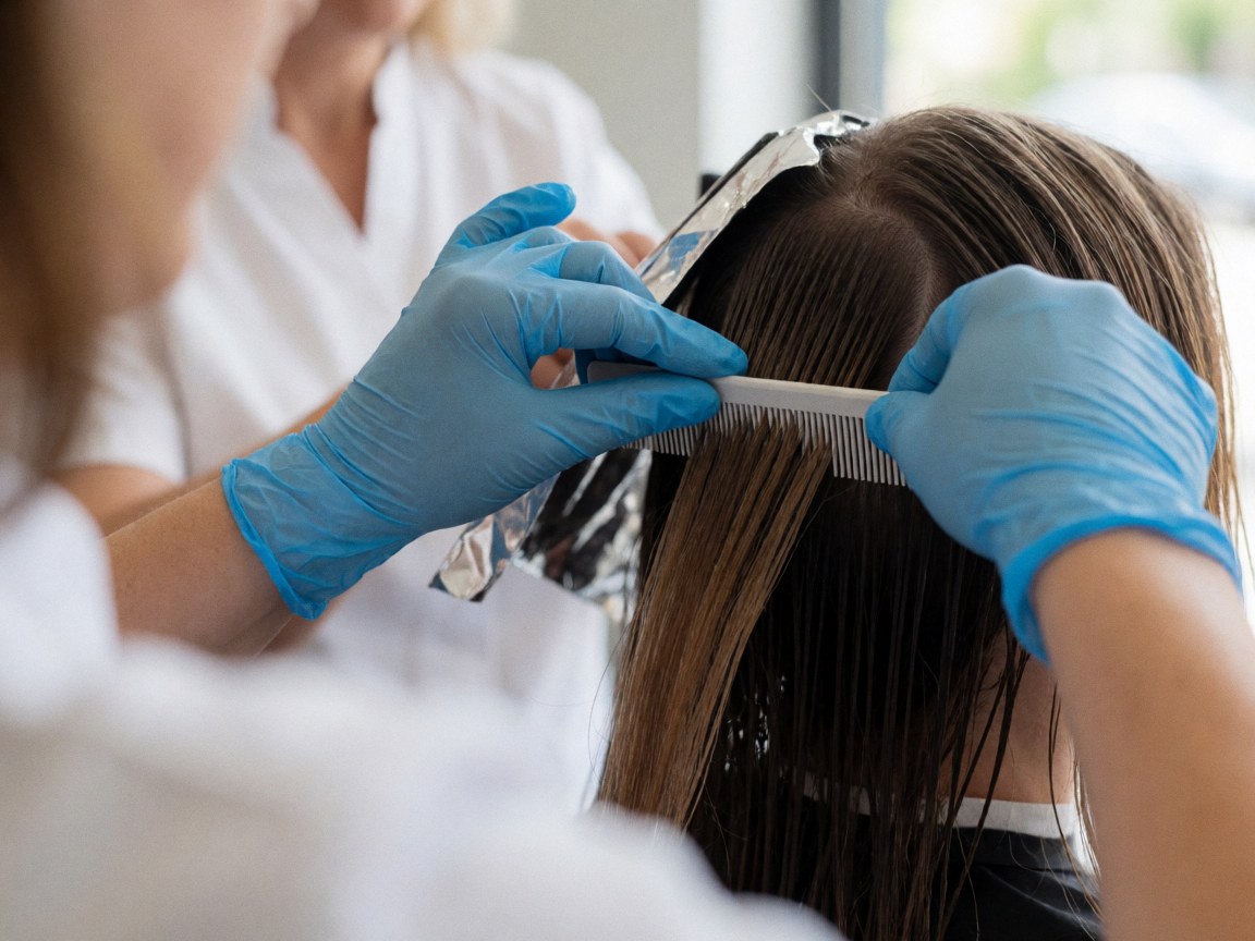 Hands wearing blue gloves sectioning wet hair with a rat-tail comb over metal foil in a salon