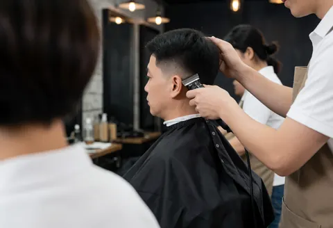 A barber uses a trimmer to cut a client's hair in a salon with industrial lights and dark walls.