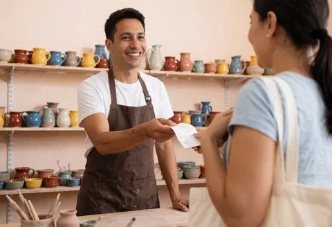 A ceramic seller hands a receipt to a customer in a workshop with colorful pottery on shelves and a tote bag.