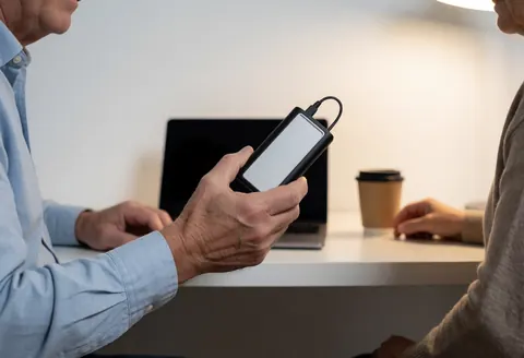 Close-up of a hand holding a connected external hard drive with a laptop and coffee cup in the background