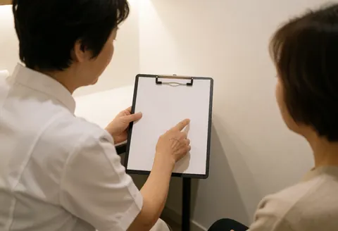 Professional in white coat points to guide on board while explaining care to seated client in consultation with white wall and warm light.