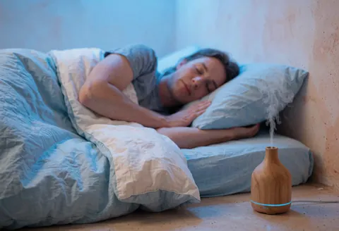 An adult sleeps peacefully in bed beside an aromatherapy diffuser emitting vapor, featuring terracotta plaster walls and soft blue lighting.