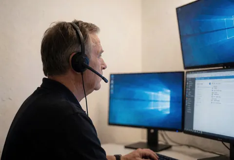 Security operator wearing a headset working at a desk with three monitors in a professional control room with light walls.