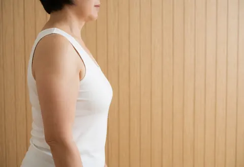 A woman in profile wearing a white blouse standing against a wooden wall with a clean background and natural light