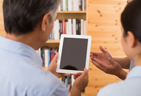 A professional and a client are reviewing information on a tablet in an office setting with a wooden bookshelf in the background.