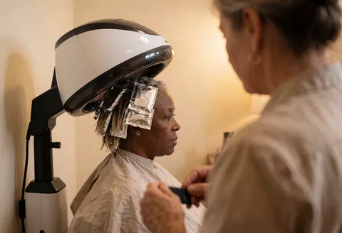 Client with foils in hair under a dryer, stylist working in a salon with beige walls and warm light