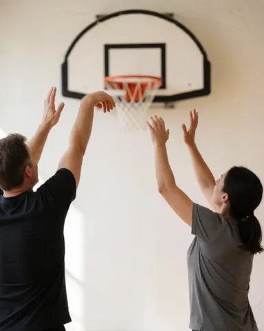 Two individuals playing basketball in a gym with white walls and natural light, showing the basketball hoop.