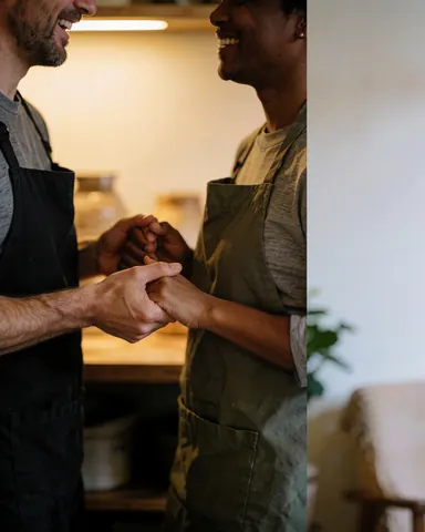 Two people wearing aprons smiling and holding hands in a warmly lit kitchen, illustrating teamwork in a small business setting.