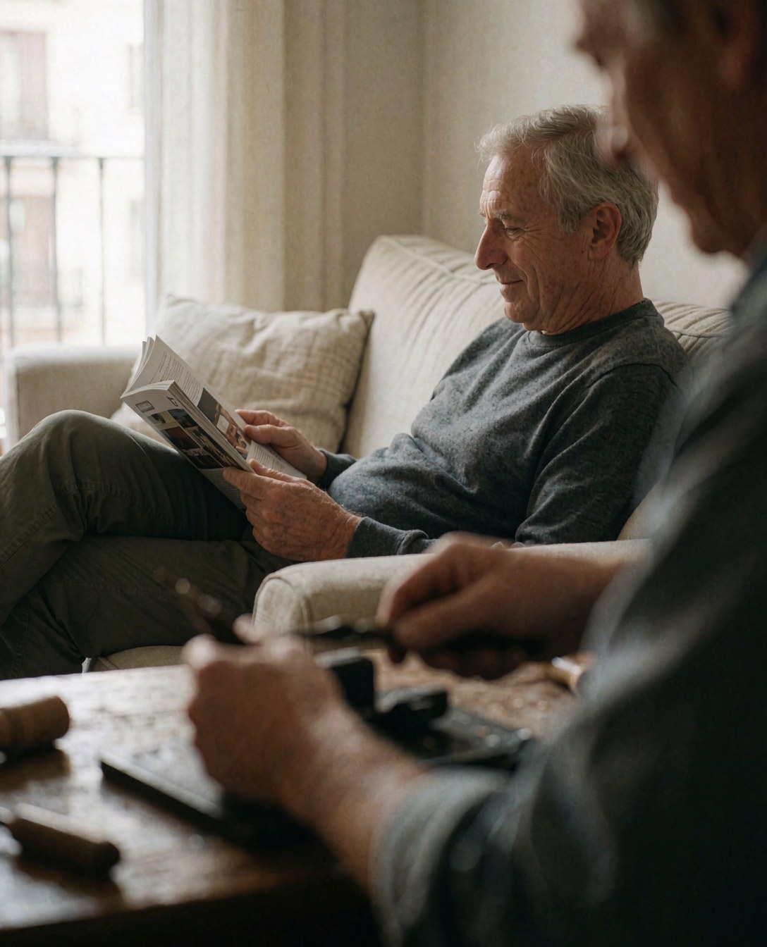 A man reads a magazine on a sofa while another works with tools at a table in a quiet, natural indoor setting.