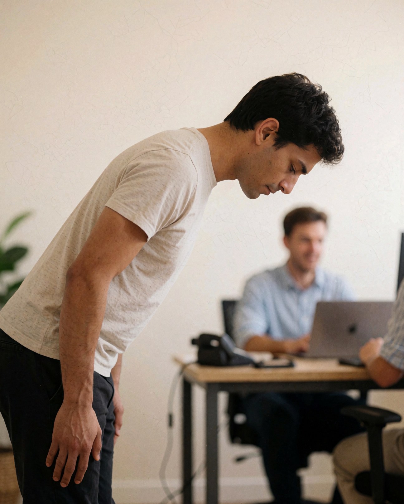 A man standing and greeting in a modern office with a colleague working on a laptop in the background.