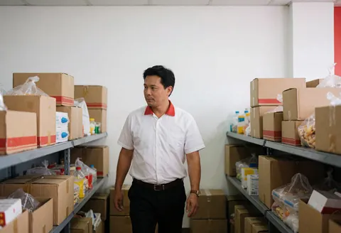 A manager walks through aisles of stacked boxes and packaged goods in a fast food storage area with white walls and practical lighting.