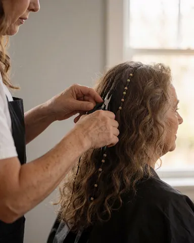 A stylist uses tweezers to apply hair extensions to a seated client near a window with natural light.