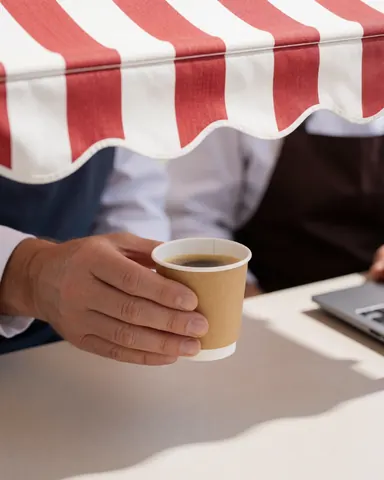 A hand holding a coffee cup under a red and white striped awning on an outdoor table, perfect for a cafe or restaurant setting.