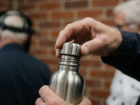 Close-up of an adult hand unscrewing the cap of a reusable metal water bottle with visible water droplets against a brick wall background.