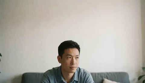A man wearing a denim shirt sits on a gray sofa, gazing to the right in a bright room with white walls and indoor plants.