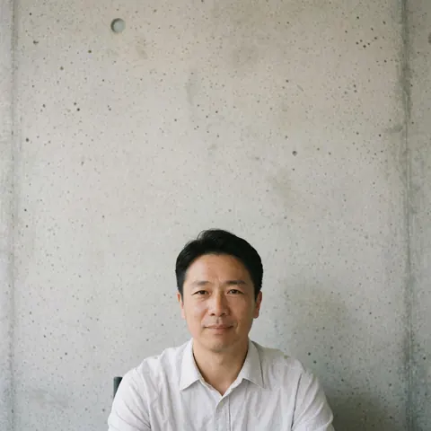 A man wearing a white shirt stands in front of a plain concrete wall with no visible business elements or background details.