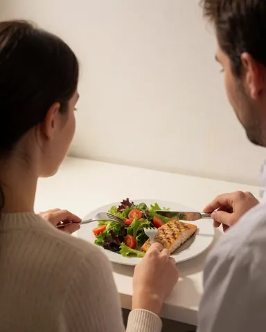 A happy client enjoys a meal of grilled fish and salad at a white table, with a fitness trainer standing in the background.