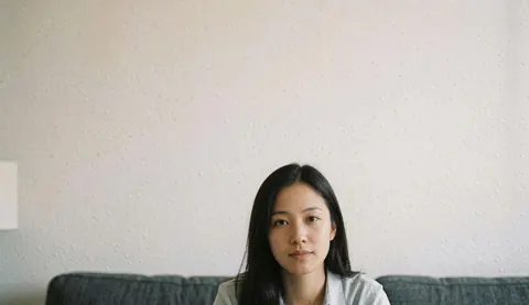 A young woman with dark hair sits on a gray sofa in a room with white textured walls, looking directly at the camera.