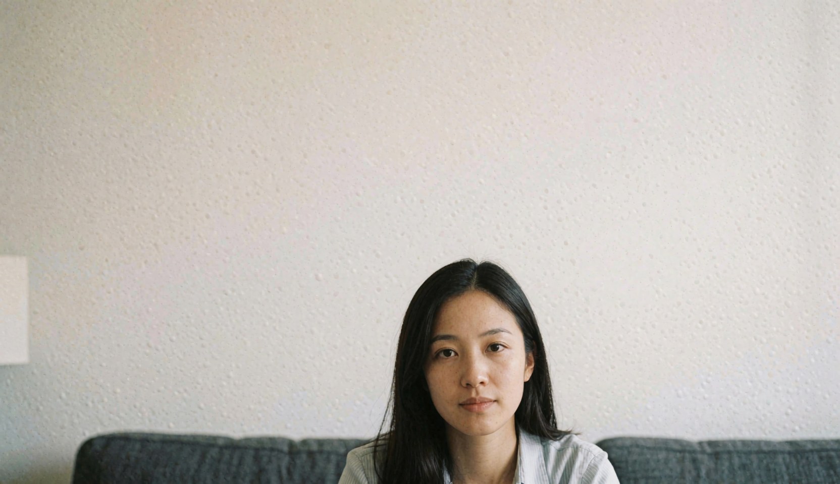 A young woman with dark hair sits on a gray sofa in a room with white textured walls, looking directly at the camera.