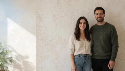 A smiling couple stands against a textured wall with natural light, suitable for business backgrounds