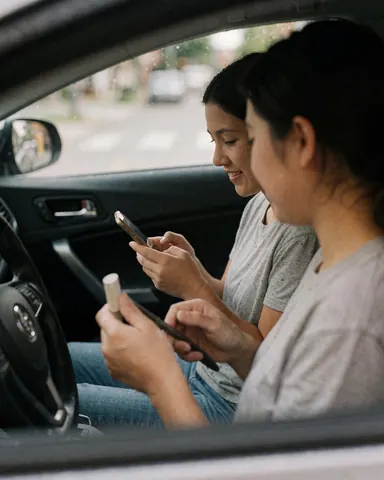 Two smiling women holding cellphones inside a parked car with the vehicle interior visible and a blurred street in the background.