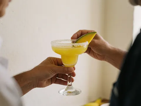 Margarita glass with salted rim and mango garnish held by hands against warm white wall