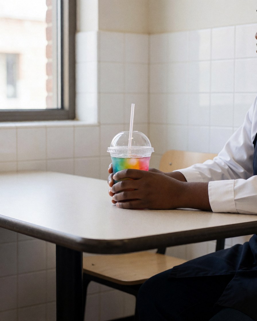 A customer enjoying a layered drink with a lid and straw at a laminated table in a colorful indoor amusement center with white tiled walls and natural window light