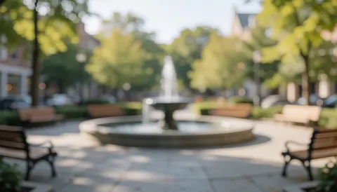 Central fountain in a plaza with wooden benches, blurred background with trees and soft light, ideal for commercial text overlays.