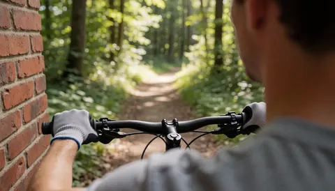 Close-up of a gloved hand gripping a bicycle handlebar while riding on a forest trail near a brick wall.